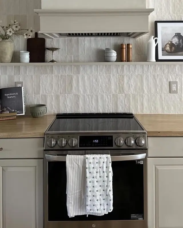 A modern kitchen features a stainless steel oven with two dish towels on the handle, a light wood countertop, and a textured backsplash with neutral tones, complemented by decorative items on the shelf above.